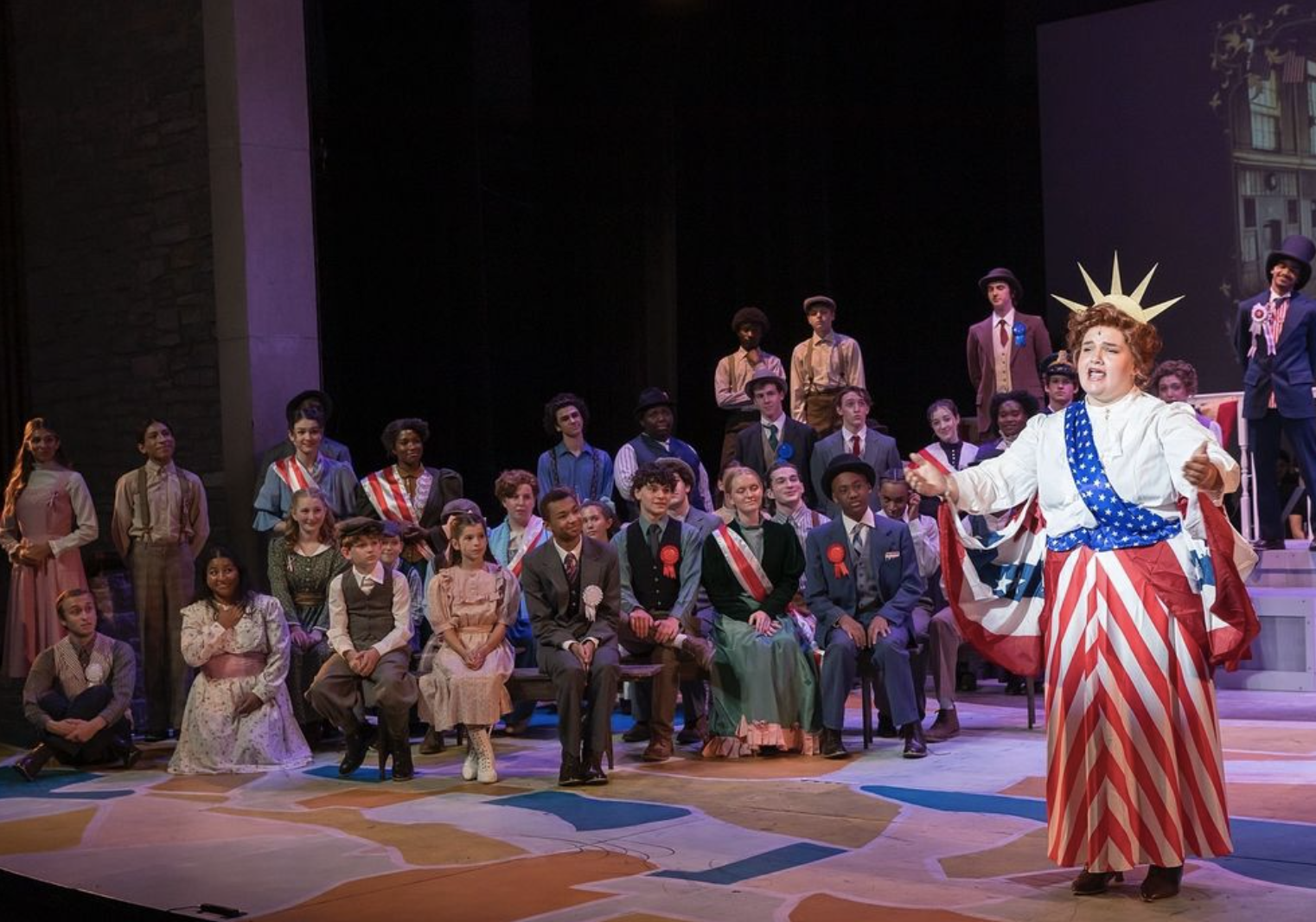 Image of the full cast of The Music Man watching a woman sing who is dressed in a white long sleeve top, red and white striped skirt, blue sash with white dots, and a liberty crown.