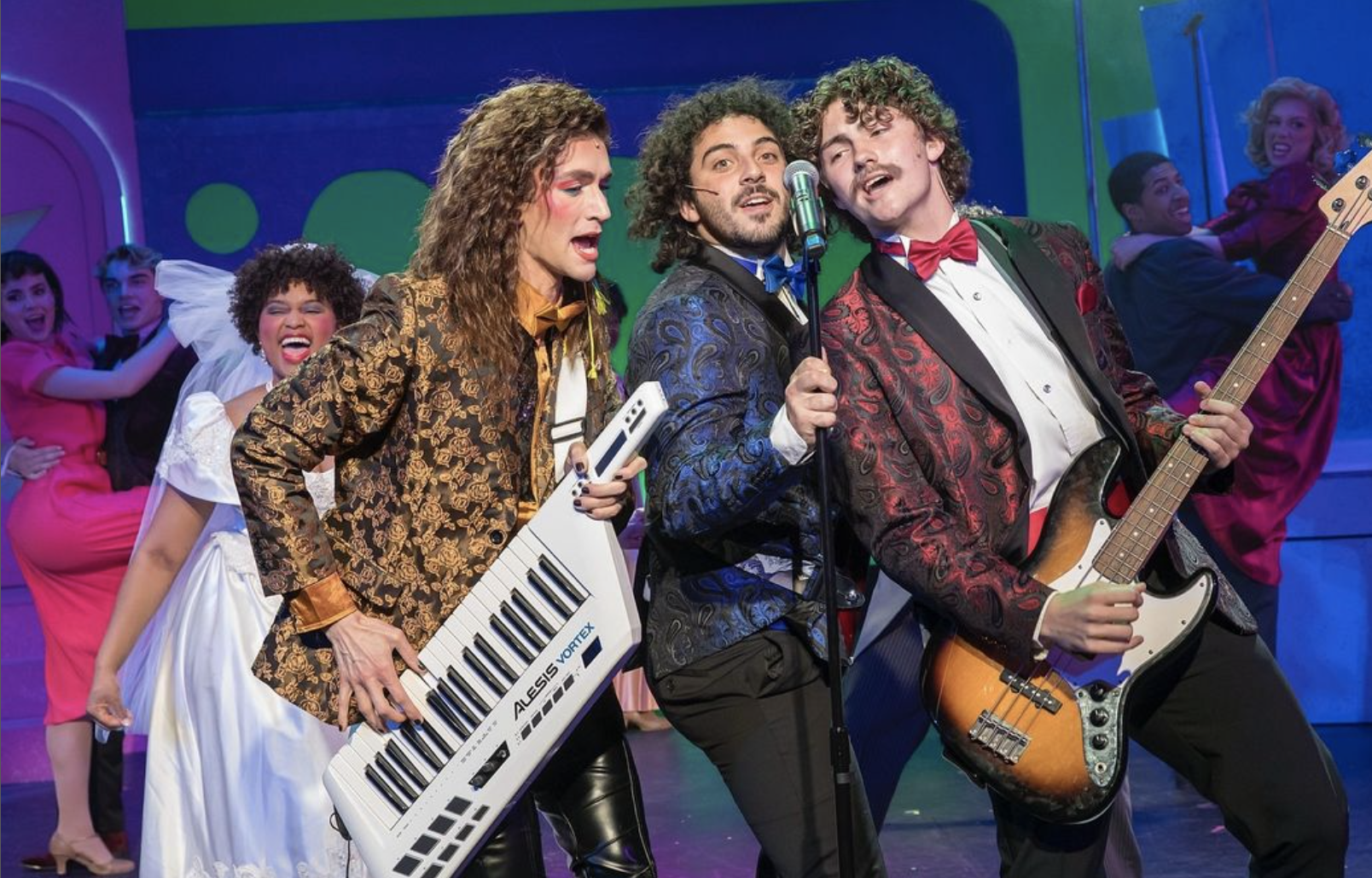 Image of three cast members of The Wedding Singer in stylish jackets singing at a microphone. One of the cast members is playing a white keyboard and one is playing an electric guitar. 